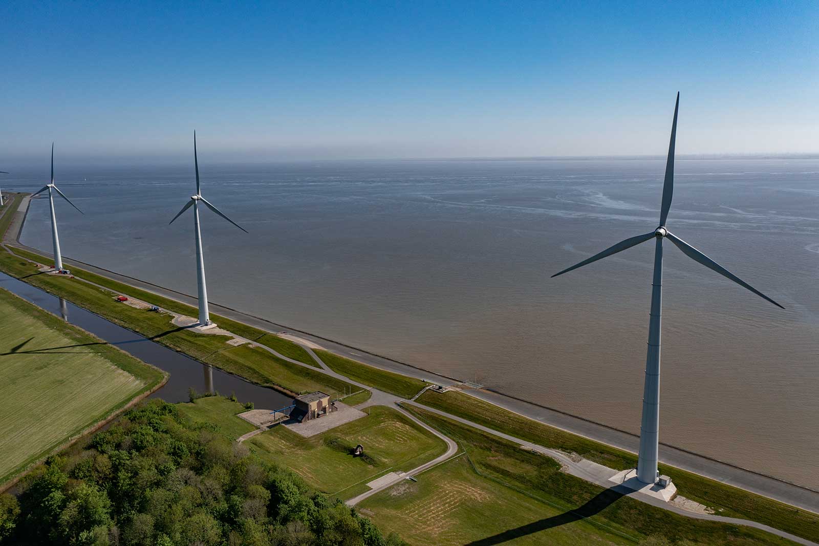 Three wind turbines by a canal, with a green field and blue sky in the background.