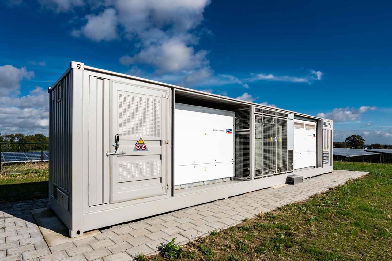 Een grote energieopslagcontainer staat op een betegeld gebied, omringd door groen gras onder een blauwe lucht met verspreide wolken.