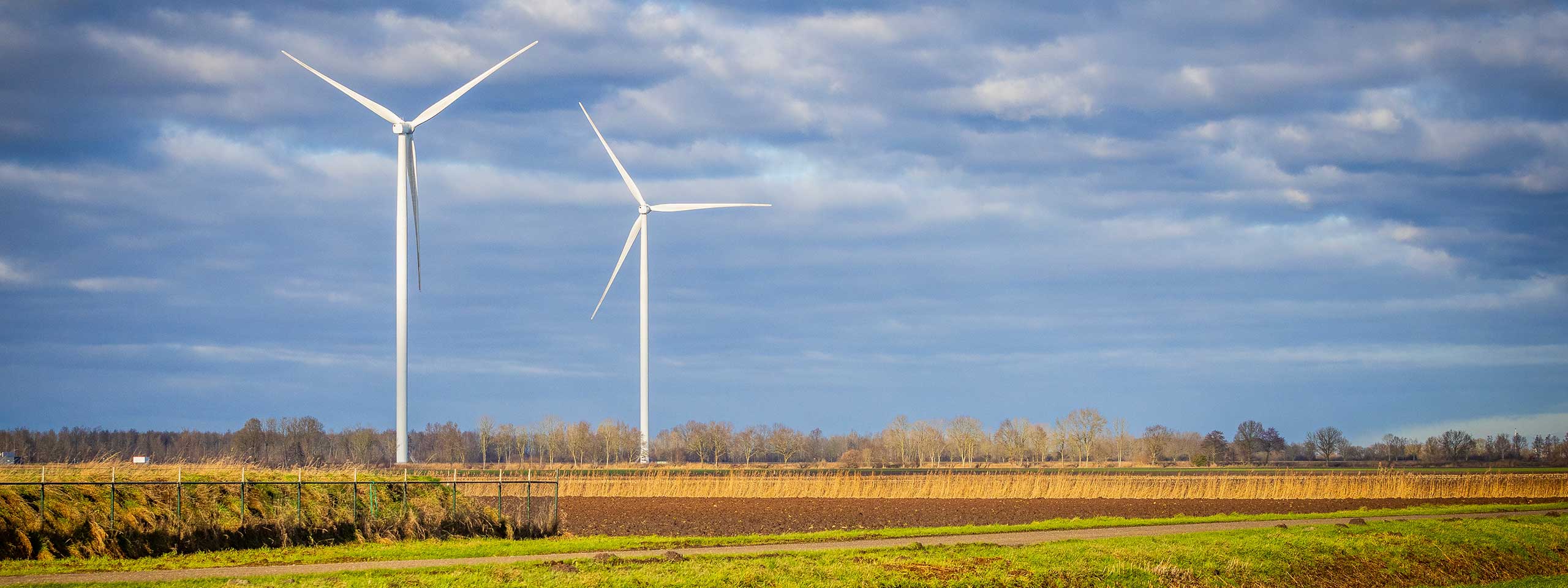 Twee grote windturbines staan hoog tegen een bewolkte lucht boven een groen veld en bruine akkers.