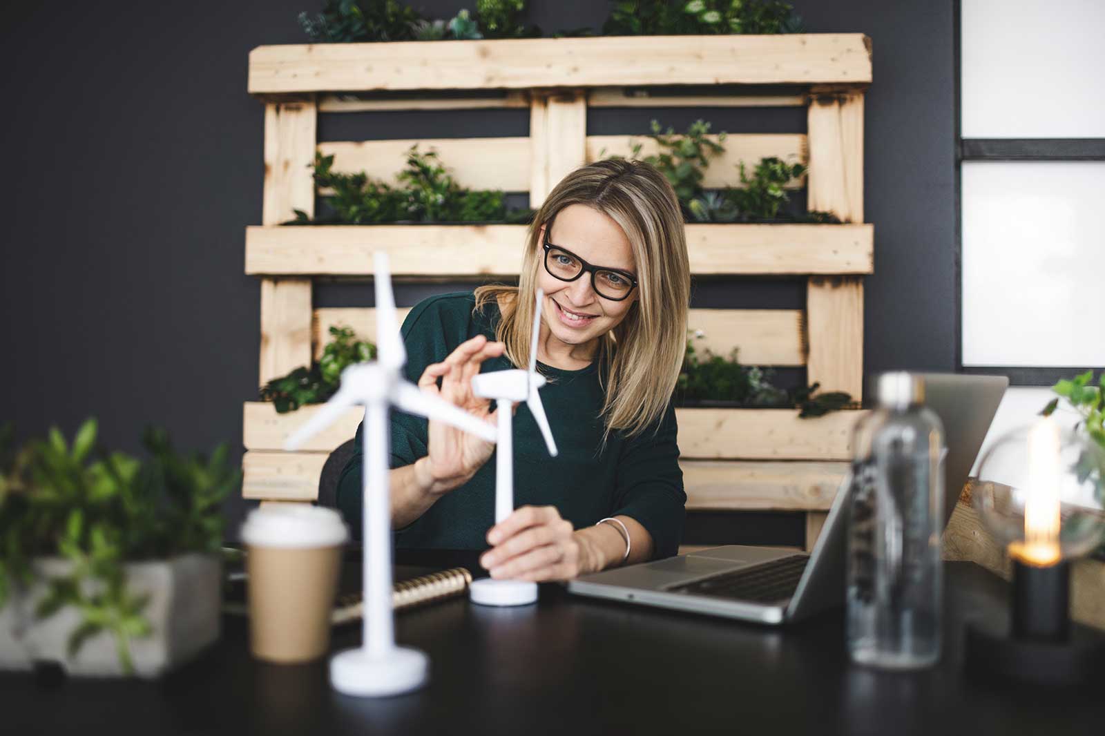 Een vrouw werkt aan een bureau met een laptop, kleine windturbines en groene planten in een moderne werkomgeving.