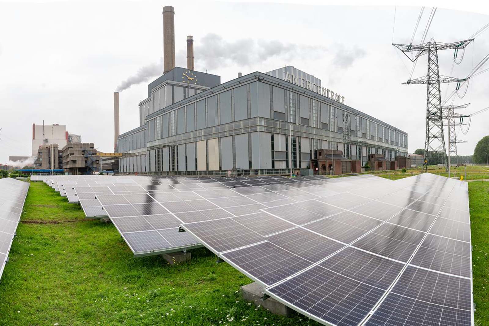 Een zonnepaneelarray voor een industriegebouw met schoorstenen, onder een bewolkte lucht, die een mix van duurzame energie en industrie toont.