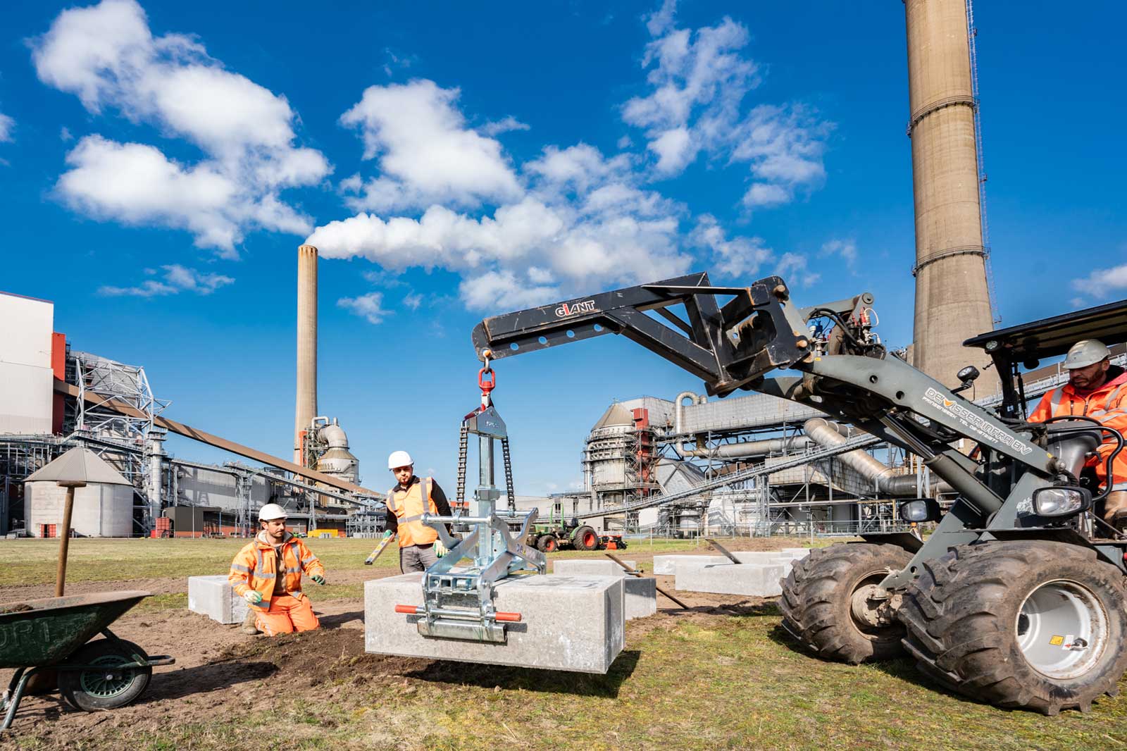 Twee arbeiders op een bouwplaats gebruiken een machine om een groot betonblok op te tillen, met industriële gebouwen op de achtergrond.