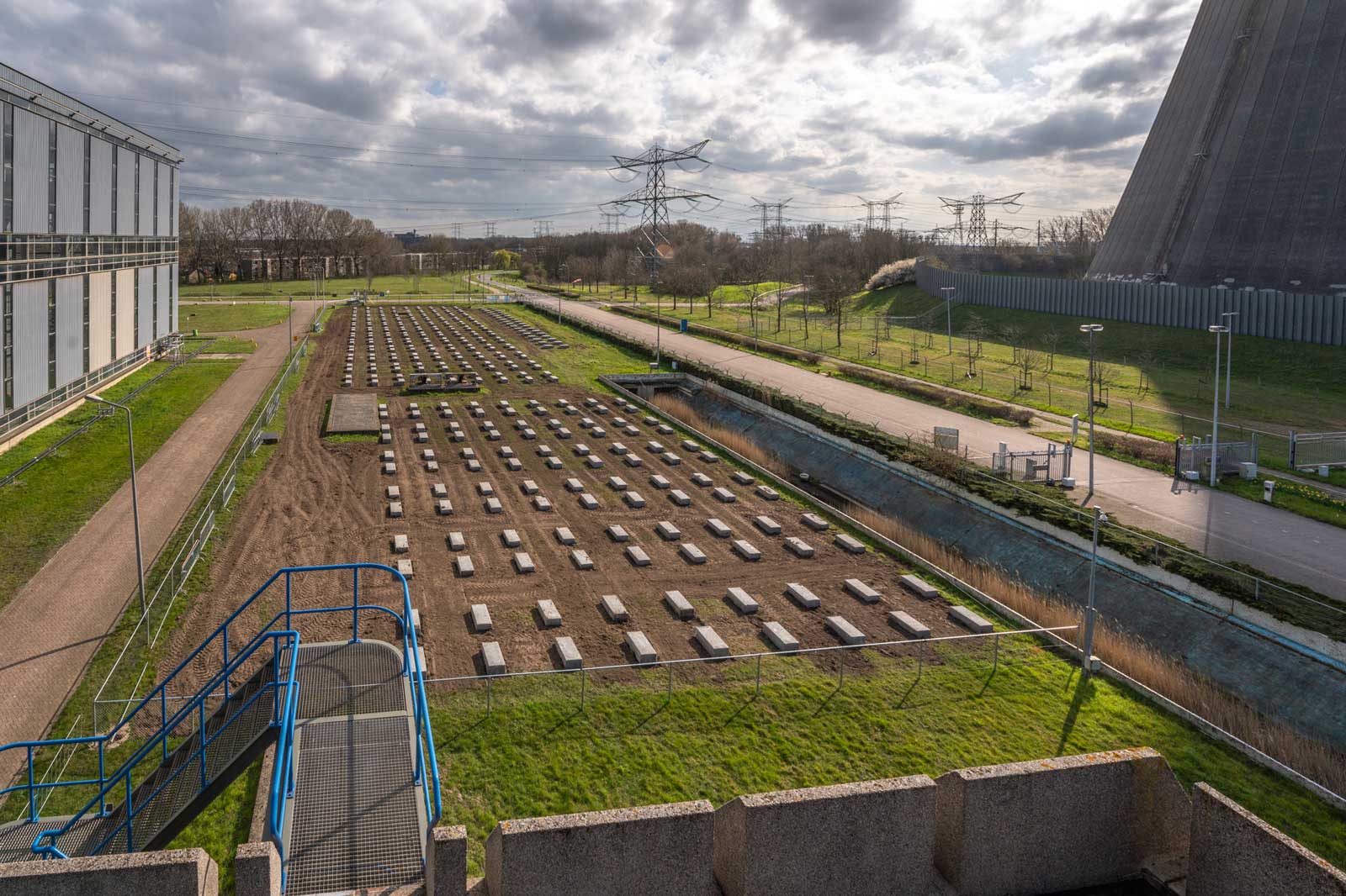 Een overzicht van een grasgebied met rijen rechthoekige percelen, een gebouw aan de linkerkant en elektriciteitsleidingen op de achtergrond.