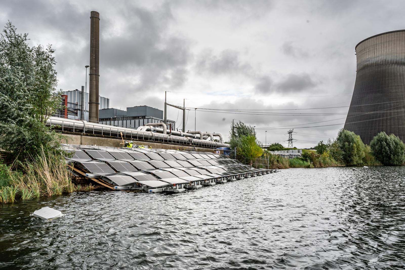 Een rij zonnepanelen op water nabij een industriële omgeving met een koeltoren en bewolkte lucht.
