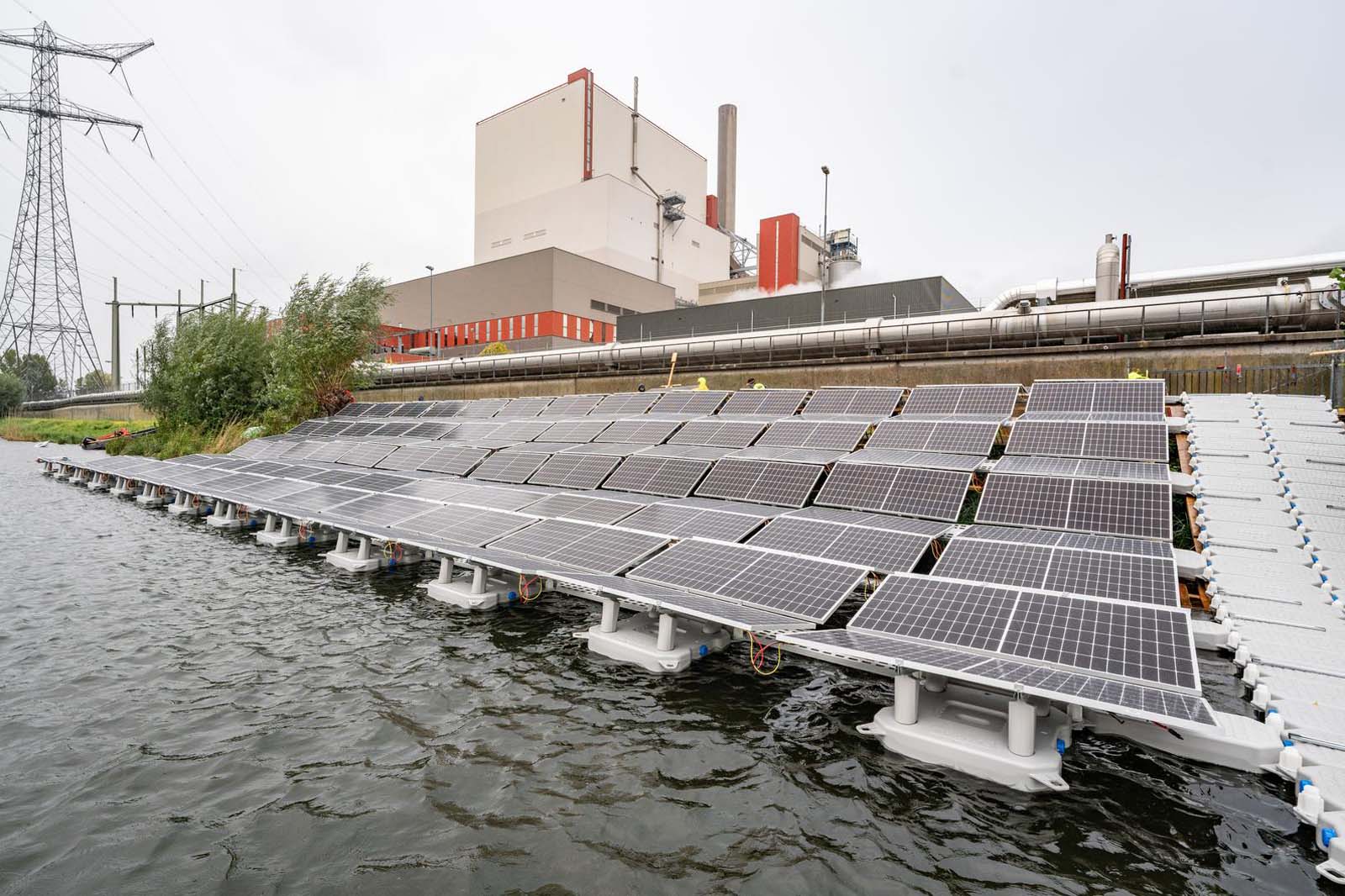 Een drijvende installatie van zonnepanelen op water, met een energiecentrale op de achtergrond en hoogspanningslijnen in de verte.