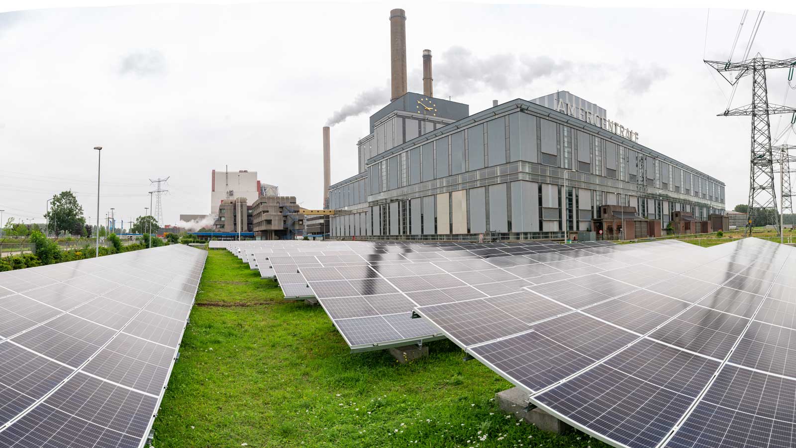 Een zonnepaneelveld voor een industrieel gebouw met rookpluimen en stroomkabels onder een bewolkte lucht.