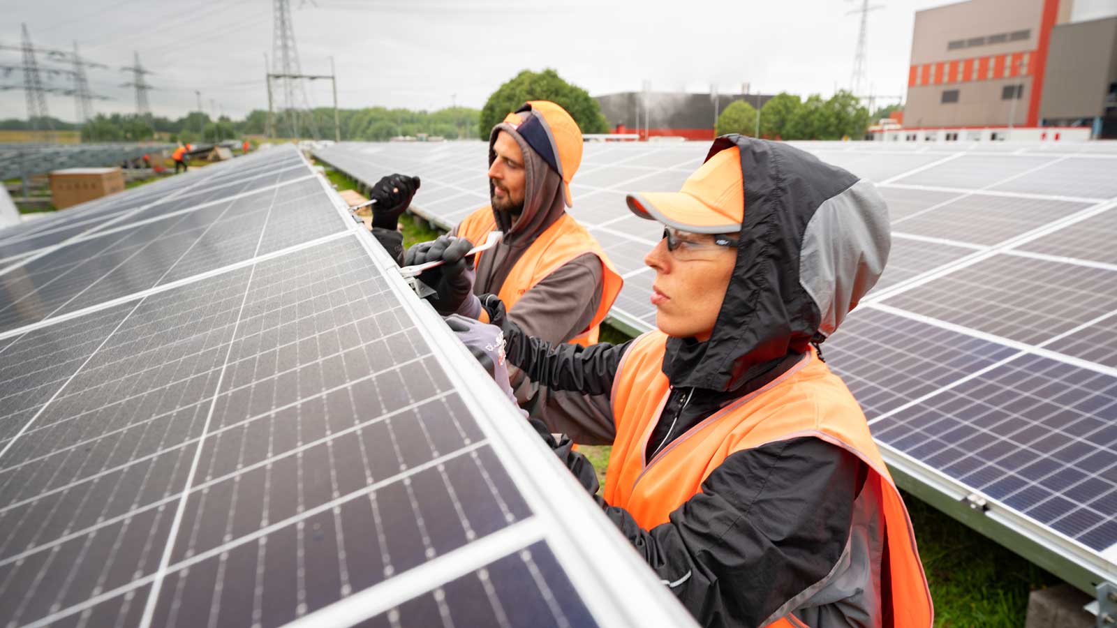 Werkers in oranje vesten installeren zonnepanelen in een veld onder een bewolkte lucht.