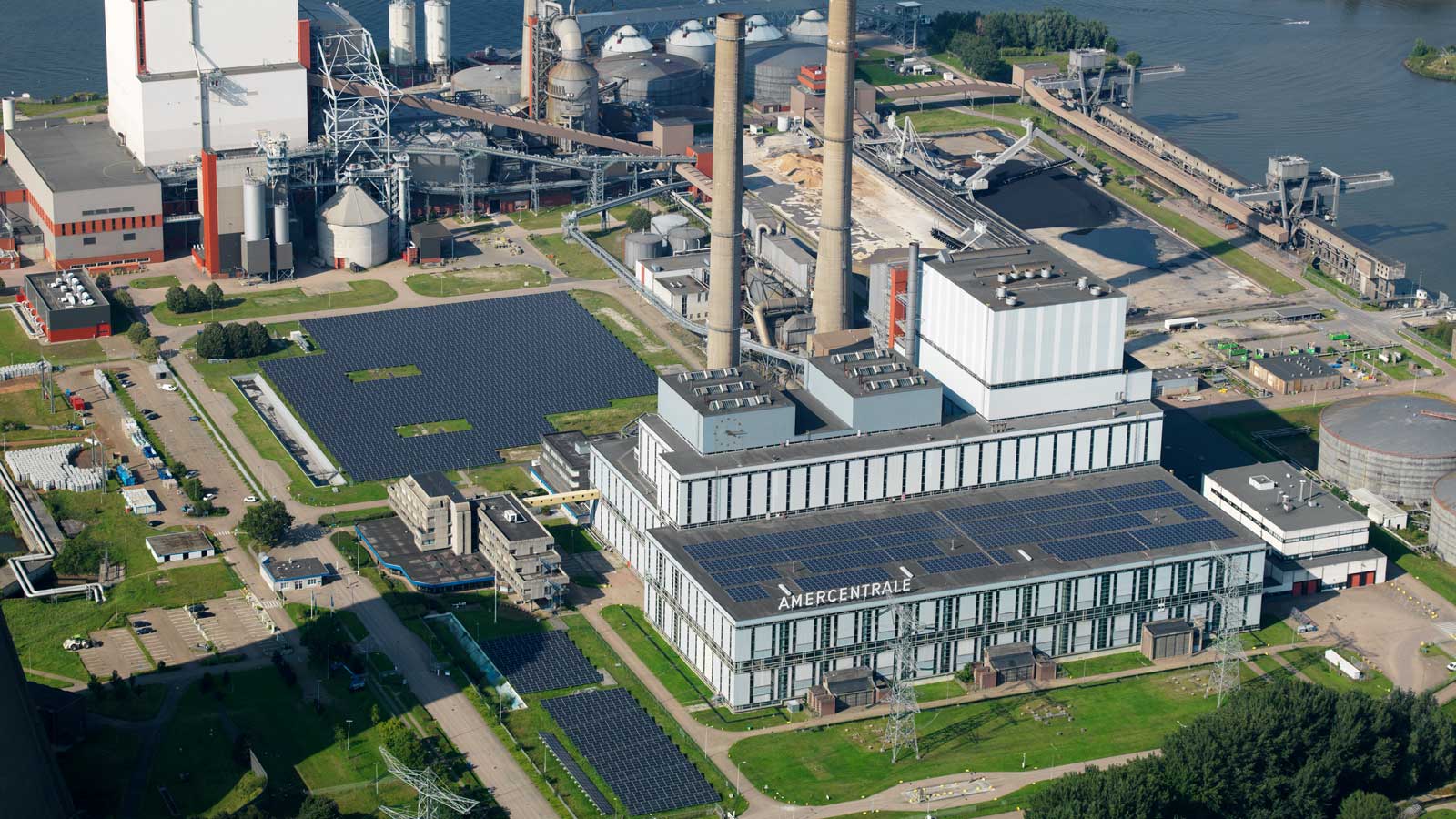 Luchtfoto van een grote elektriciteitscentrale met zonnepanelen, hoge schoorstenen en omliggende industriële faciliteiten nabij water.