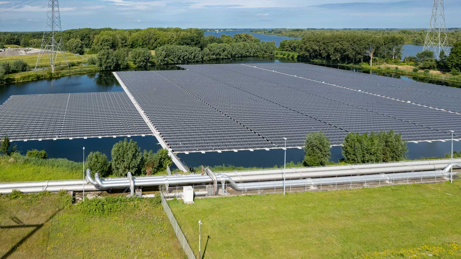 Een grote zonnepaneelinstallatie drijft op een wateroppervlak, omgeven door groen en elektriciteitsleidingen op de achtergrond.