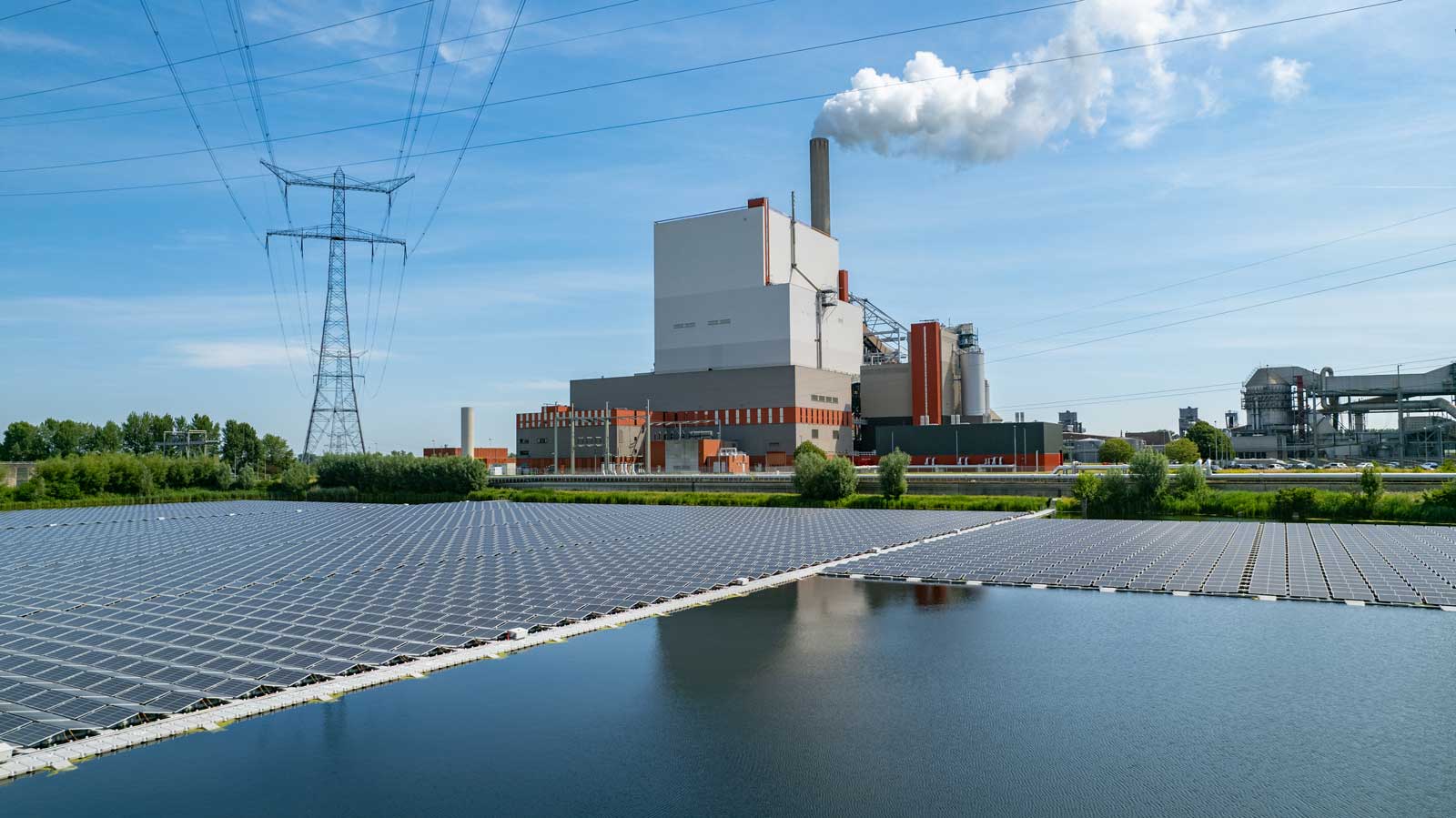 Een thermische centrale naast een zonnepaneelveld, omringd door groen onder een heldere blauwe lucht.