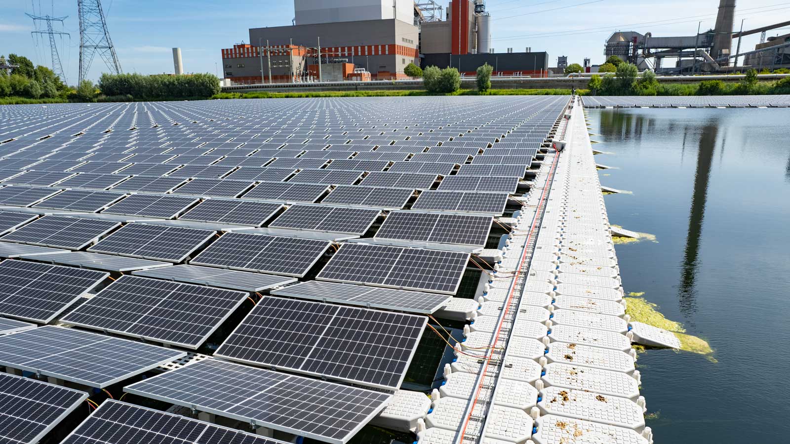 Een zonnepaneelarray drijft op water, weerspiegeld door de blauwe lucht en omringd door groen en industriële gebouwen.
