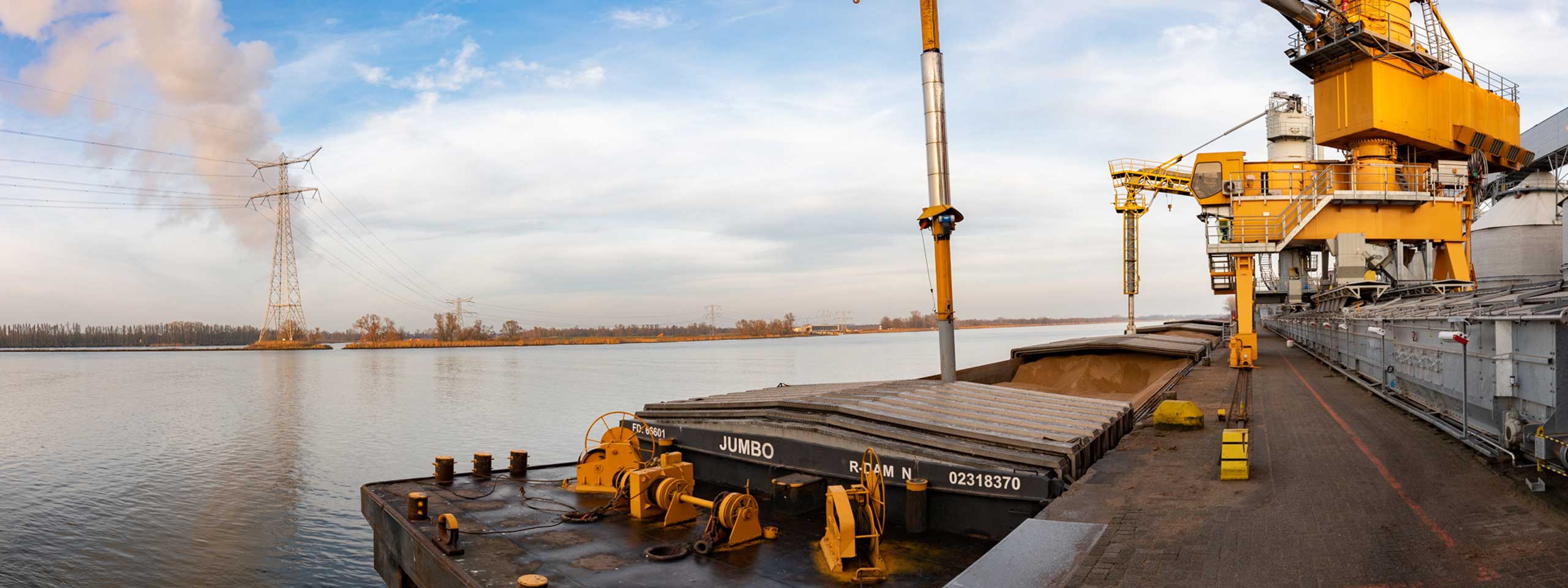 Een binnenschip geladen met materialen is aangemeerd naast een gele kraan aan de rivier, met hoogspanningslijnen en bomen op de achtergrond.