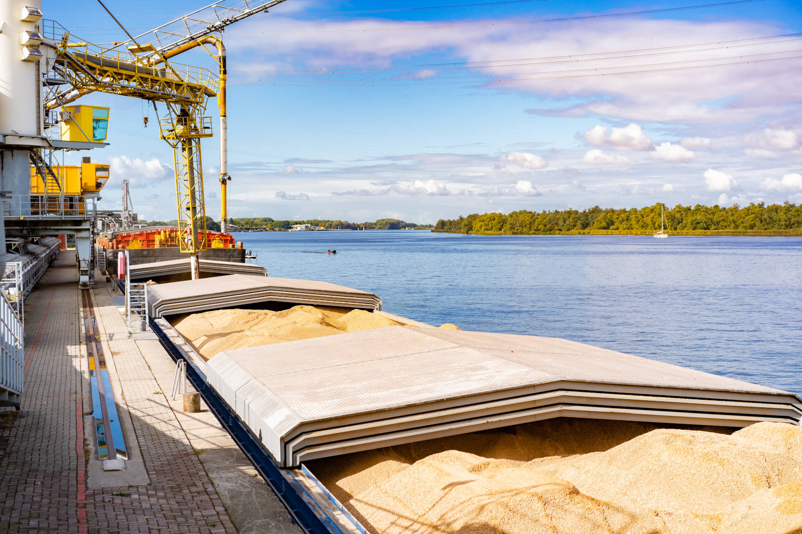 A cargo ship docked beside a river, with sand piled on the deck and cranes in the background under a partly cloudy sky.