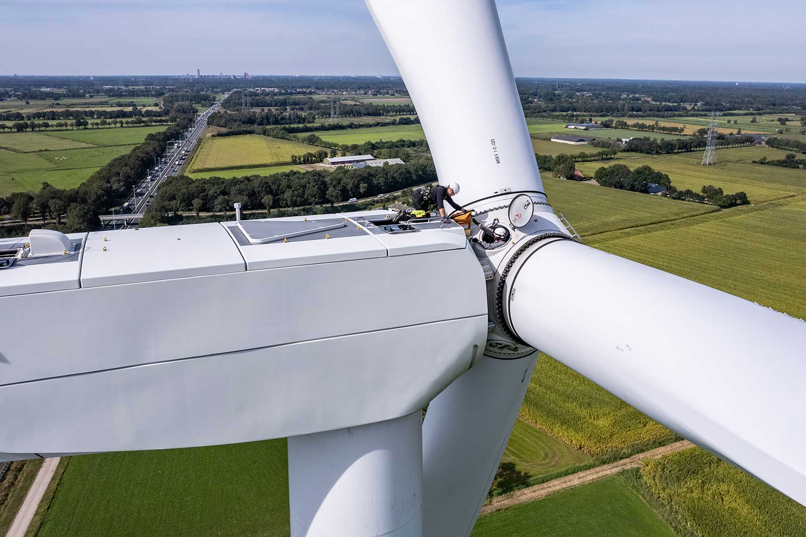 Een windturbine met een technicus die aan de rotor werkt, met uitzicht op groene velden en een snelweg.