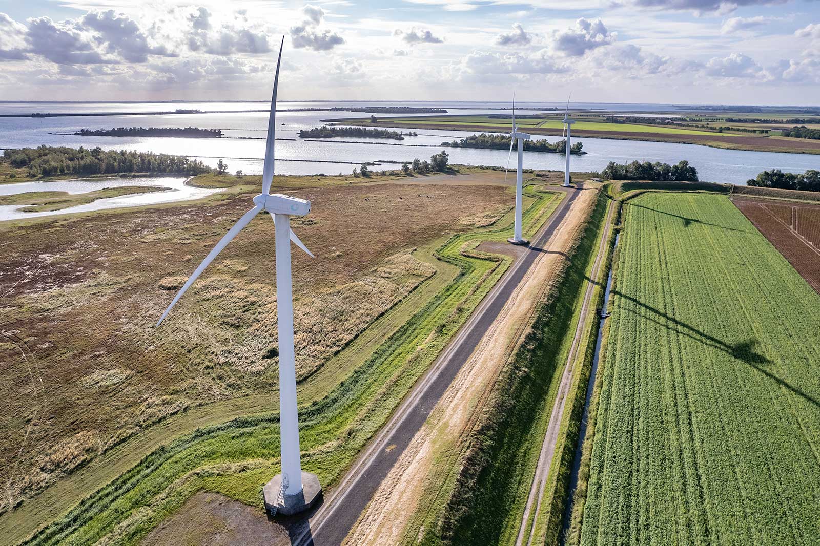 Luchtfoto van drie windturbines naast een rivier en groene velden onder een bewolkte lucht.