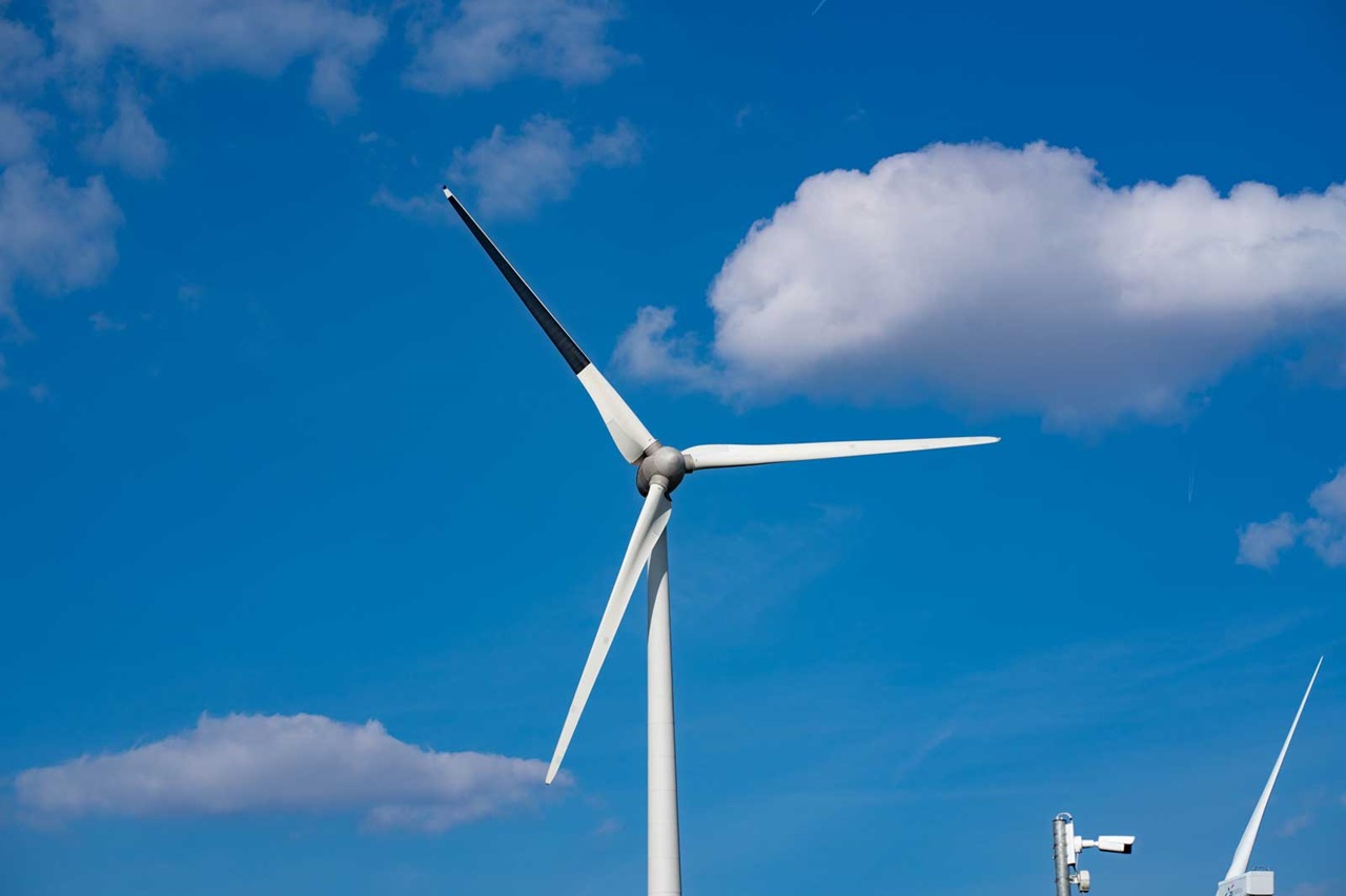 Een hoge windturbine met drie bladen tegen een blauwe lucht, met pluizige witte wolken.