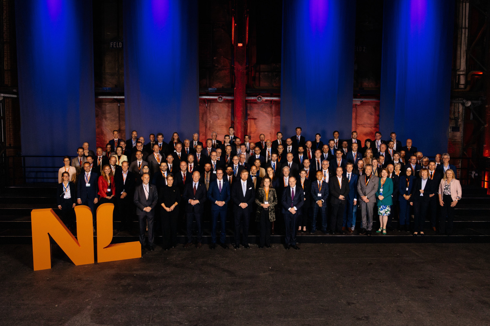 Een groep mensen in formele kleding staat voor een foto, met paarse lichten en een oranje "NL" teken op de voorgrond.