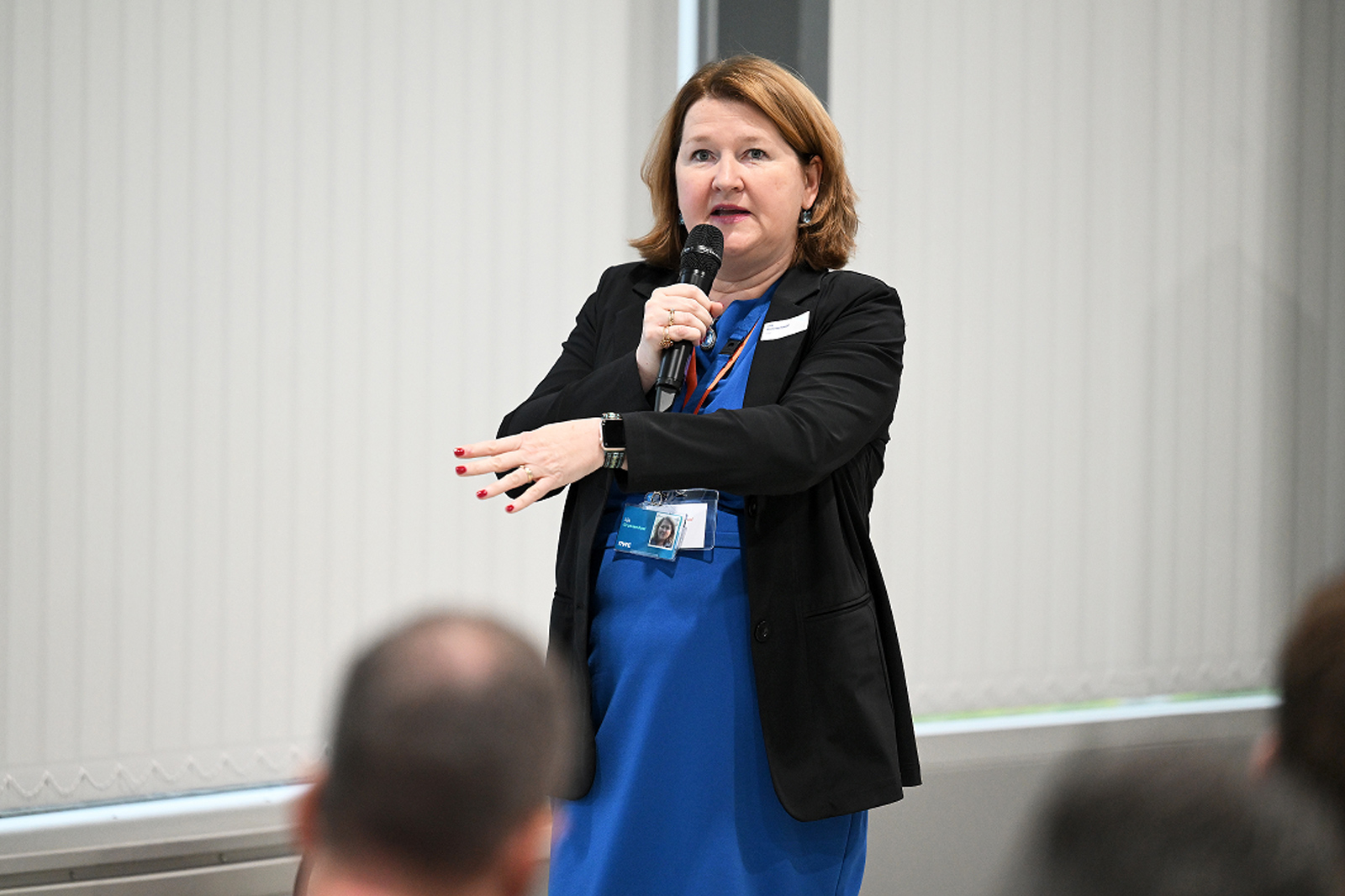 Een vrouw in een blauwe jurk en zwart jasje spreekt in een microfoon, met haar hand gebarend, in een conferentieruimte.