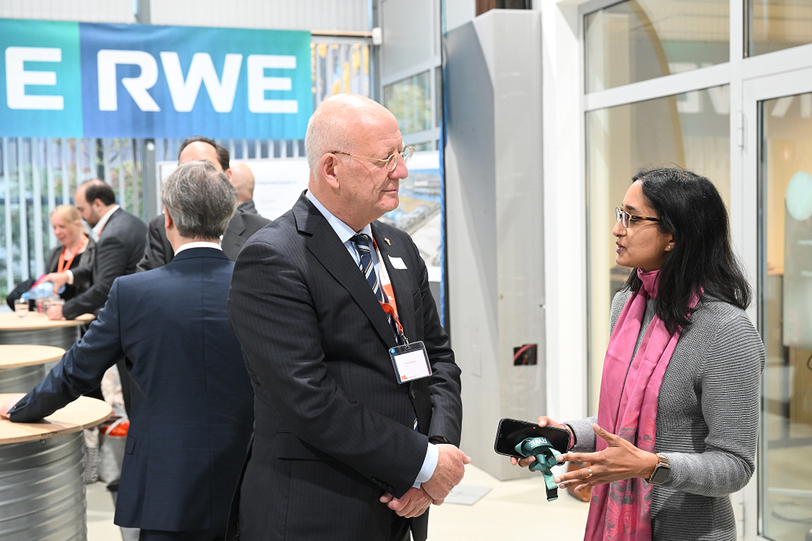 Twee professionals voeren een gesprek op een evenement, met een blauw RWE-spandoek op de achtergrond en verschillende aanwezigen die zich om hen heen bewegen.