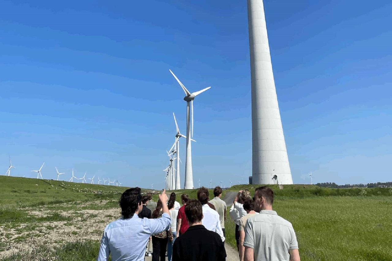 Een groep mensen die langs een pad loopt dat omringd is door grote windturbines onder een heldere blauwe lucht.
