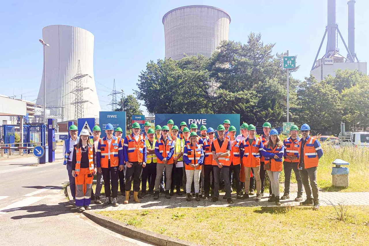 A group of workers in bright orange vests and blue helmets poses at an industrial site with cooling towers in the background.