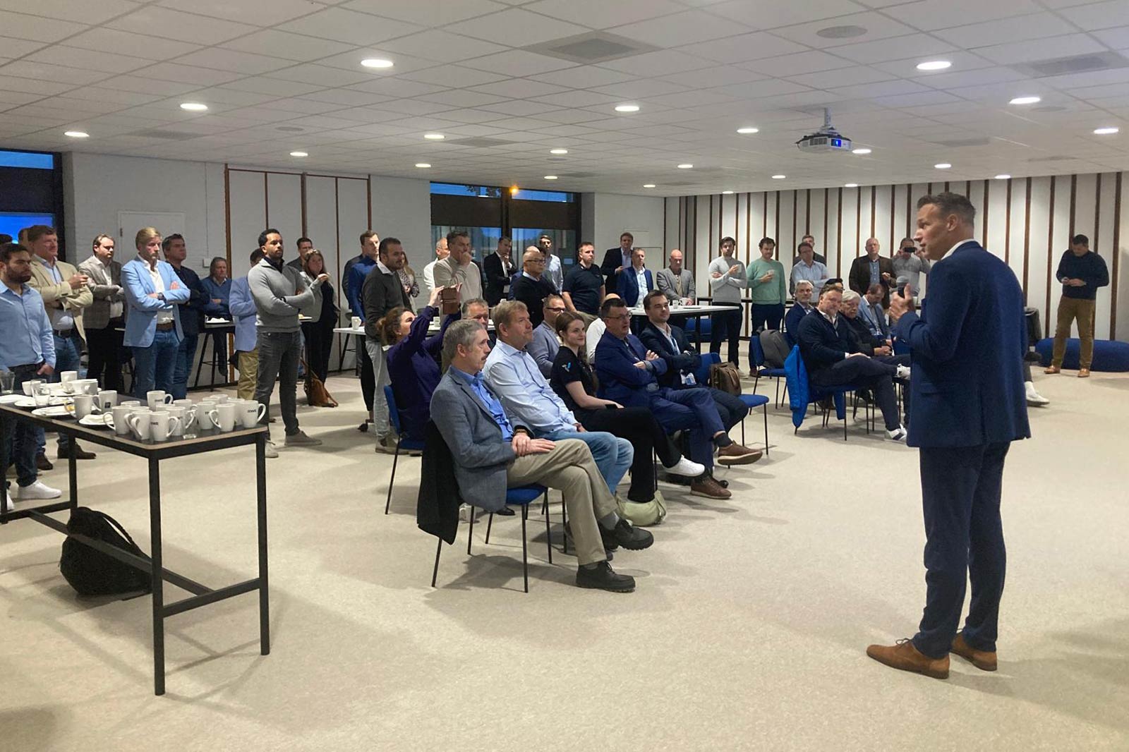 Een spreker spreekt een aandachtig publiek toe in een moderne conferentieruimte met tafels en stoelen voor een presentatie.