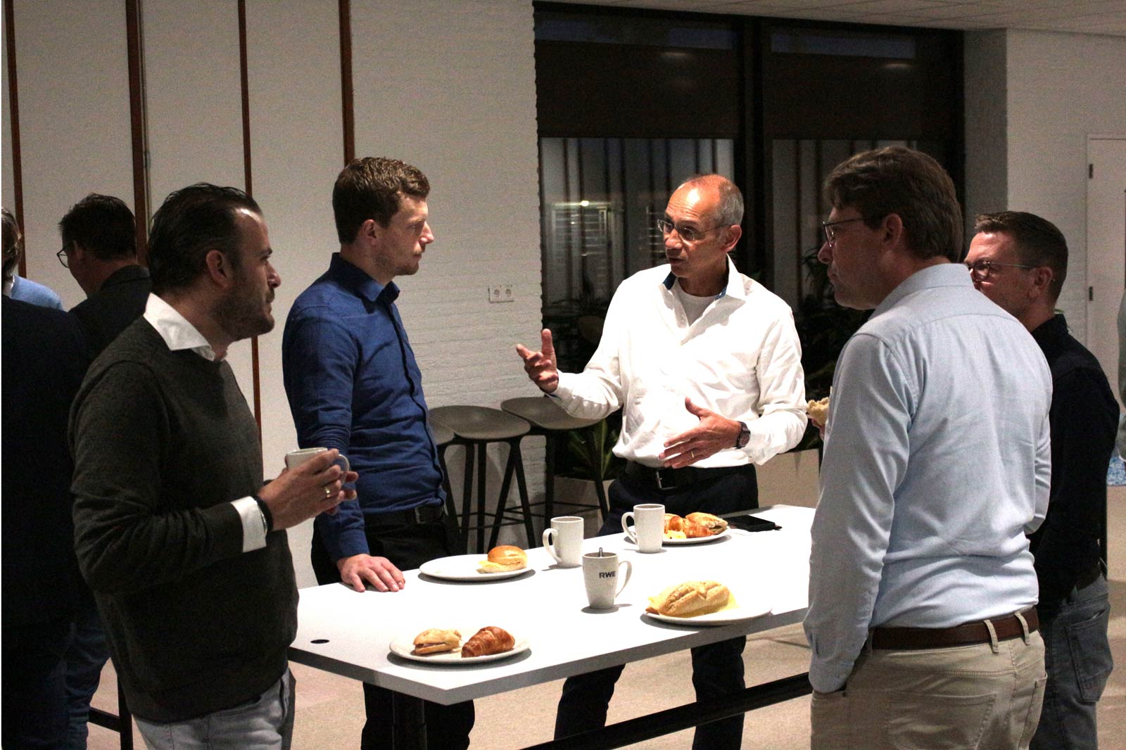 A group of professionals engaging in conversation around a table with pastries and coffee in a modern office setting.