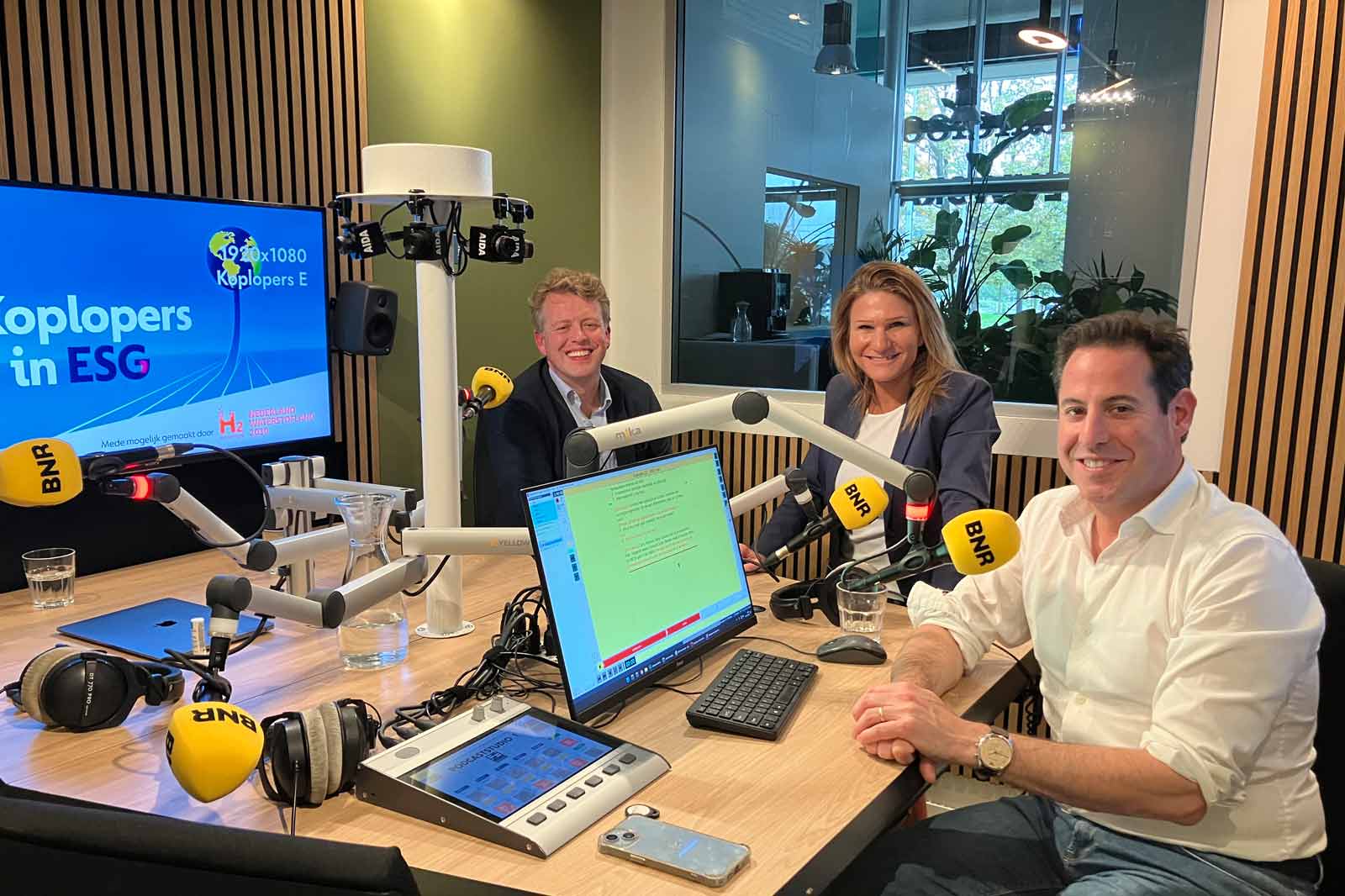 Een radiostudio met gasten die ESG-onderwerpen bespreken, met apparatuur en een presentatiescherm.