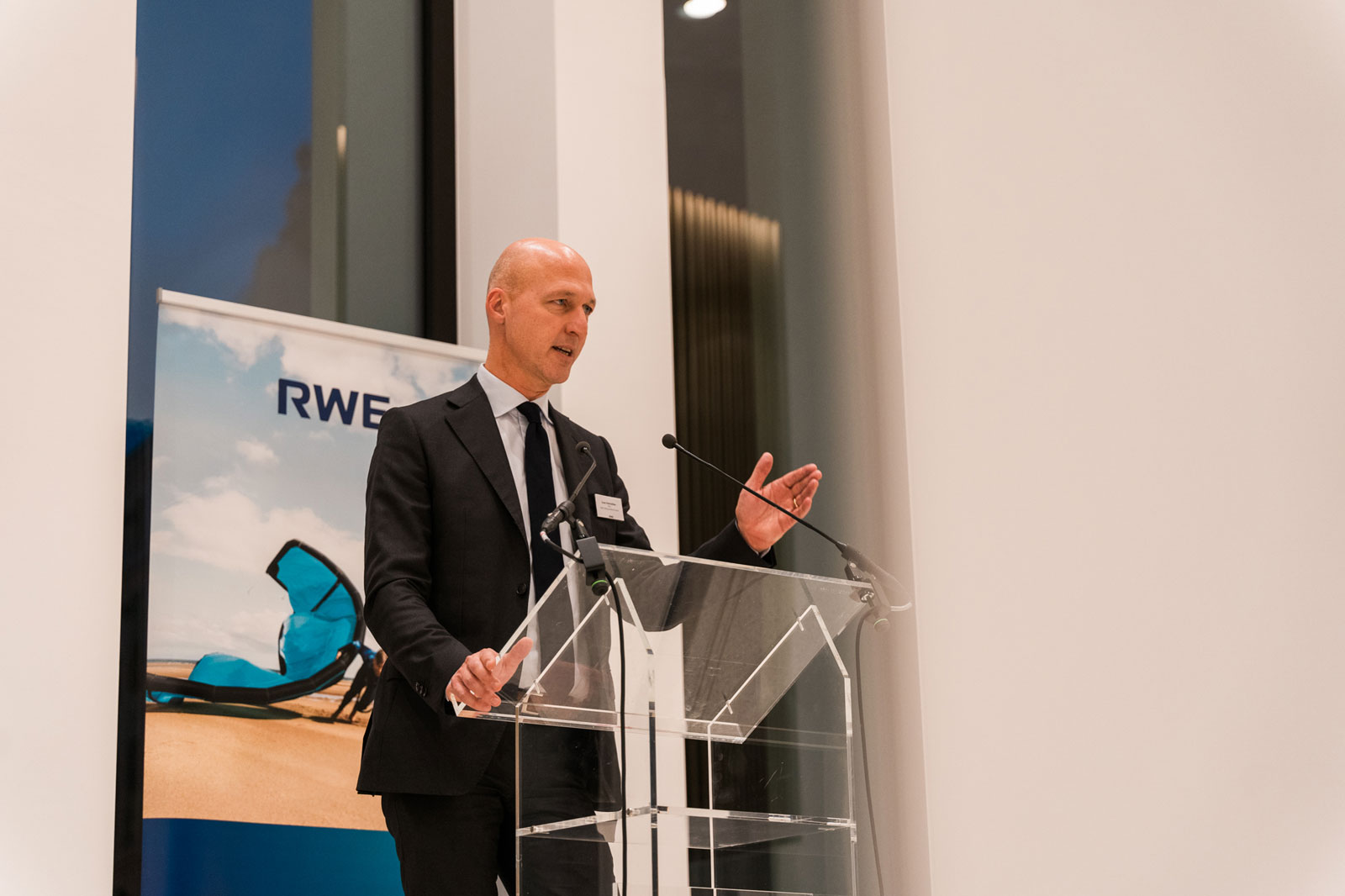 Een spreker gebaart terwijl hij presenteert op een evenement, met een grote RWE-banner op de achtergrond.
