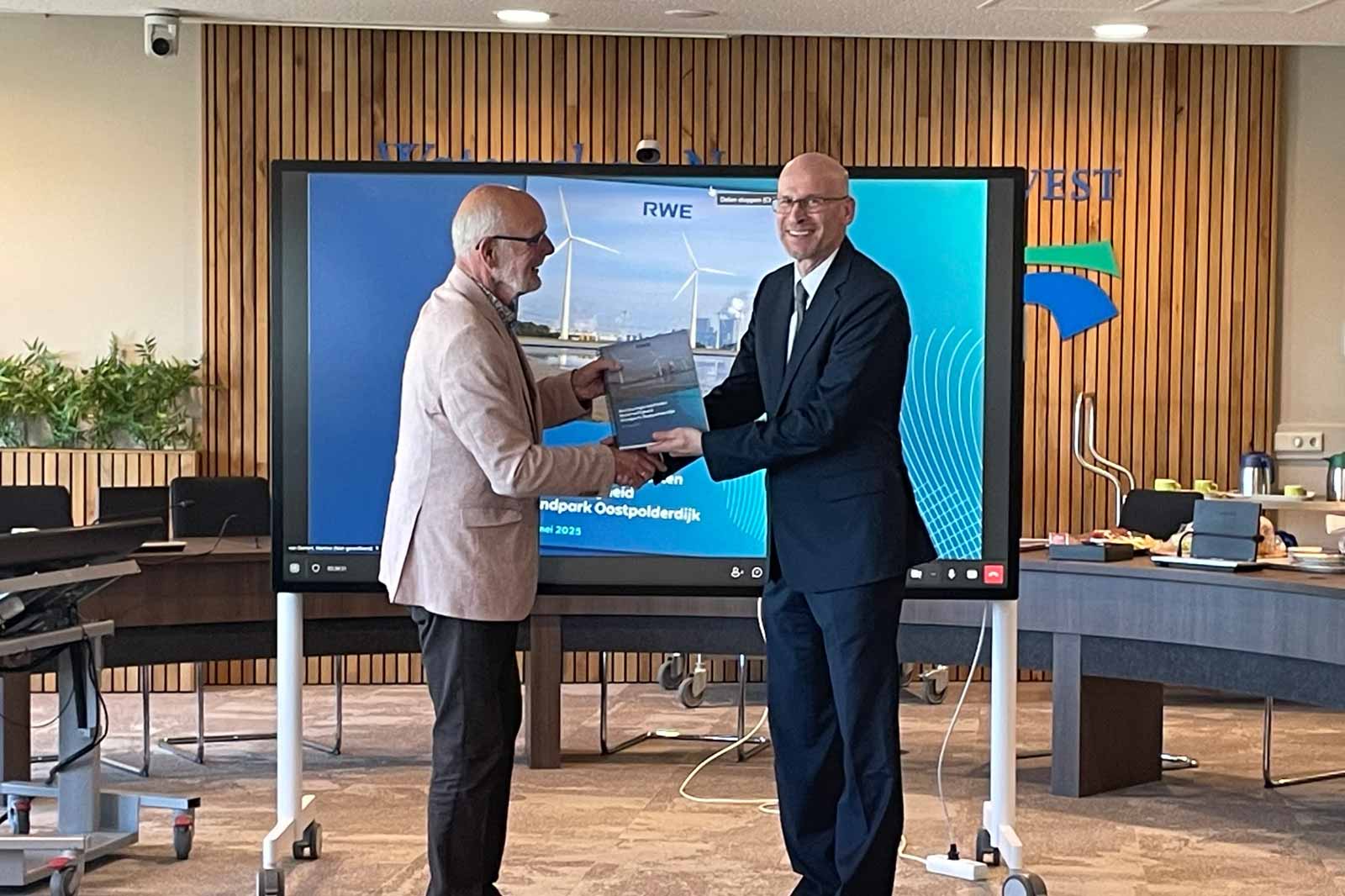 Two men exchange a document in an office setting, with a digital display of wind turbines in the background.
