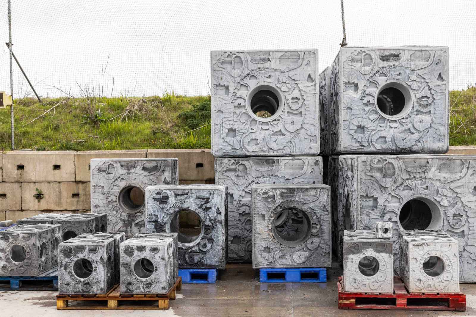 Een verzameling grote betonnen blokken met ronde gaten, gestapeld op blauwe en bruine pallets, tegen een grasachtig achtergrond.