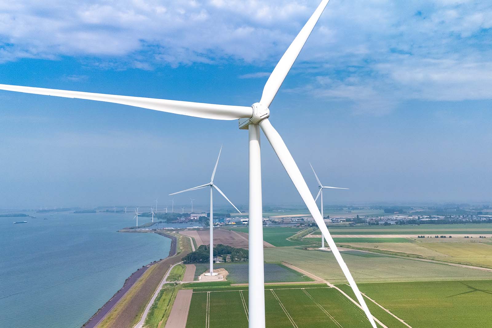 Luchtfoto van windturbines nabij een waterweg, omgeven door groene velden en een blauwe lucht.