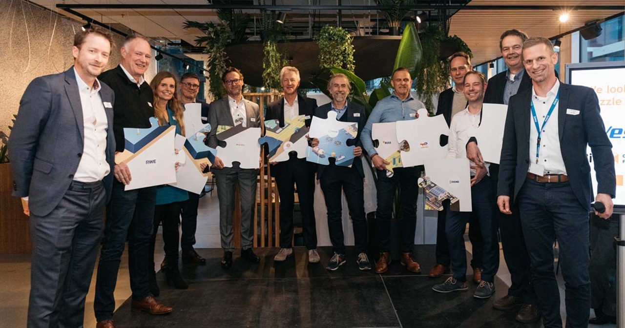 A group of people holds puzzle pieces with various designs at an indoor event surrounded by plants.