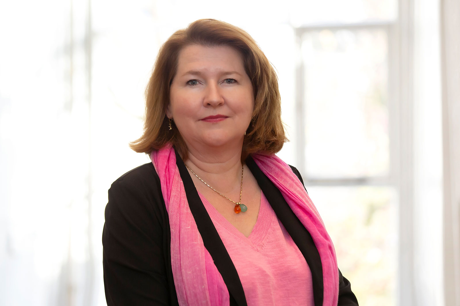 Een vrouw in een roze top met een zwarte jas en een roze sjaal glimlacht zachtjes, tegen een heldere, vage achtergrond.