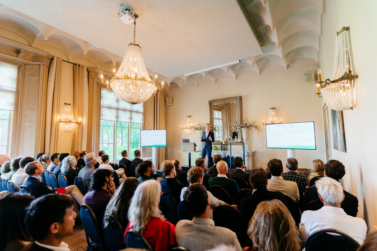 A speaker engages an audience in a stylish room with chandeliers, large windows, and a presentation screen.