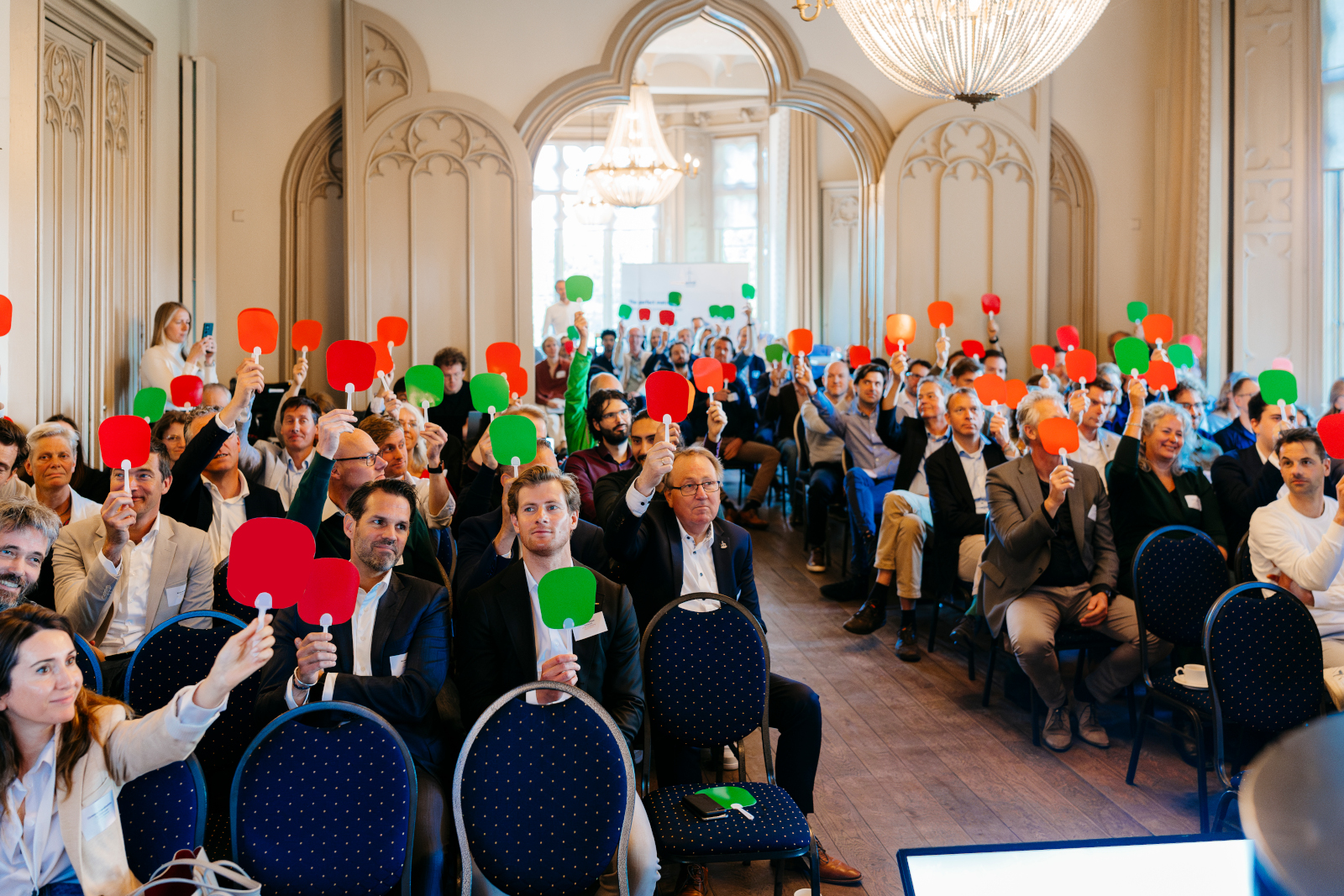 A crowd holds up red and green paddles during a meeting in an elegantly designed room, showcasing engagement and participation.