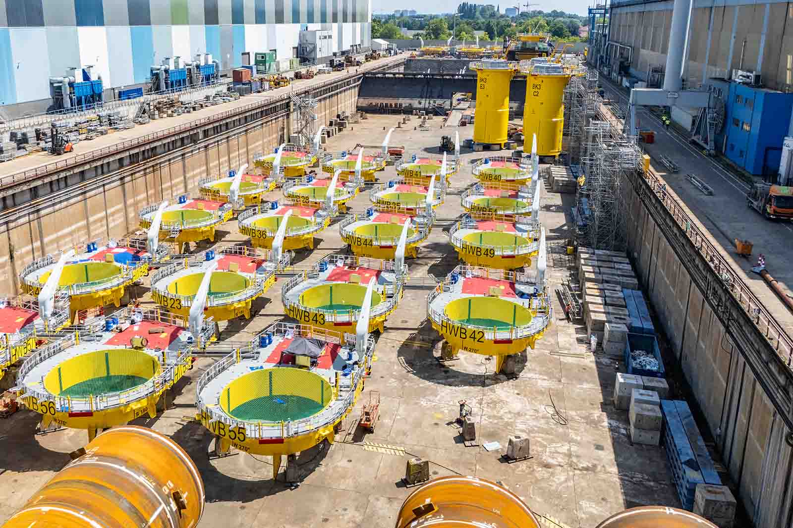Rijen gele stalen ronde drijflichamen met groene binnenbekleding en rode toegangsdeksels op een betonnen scheepswerf, één gemarkeerd "NWB42".