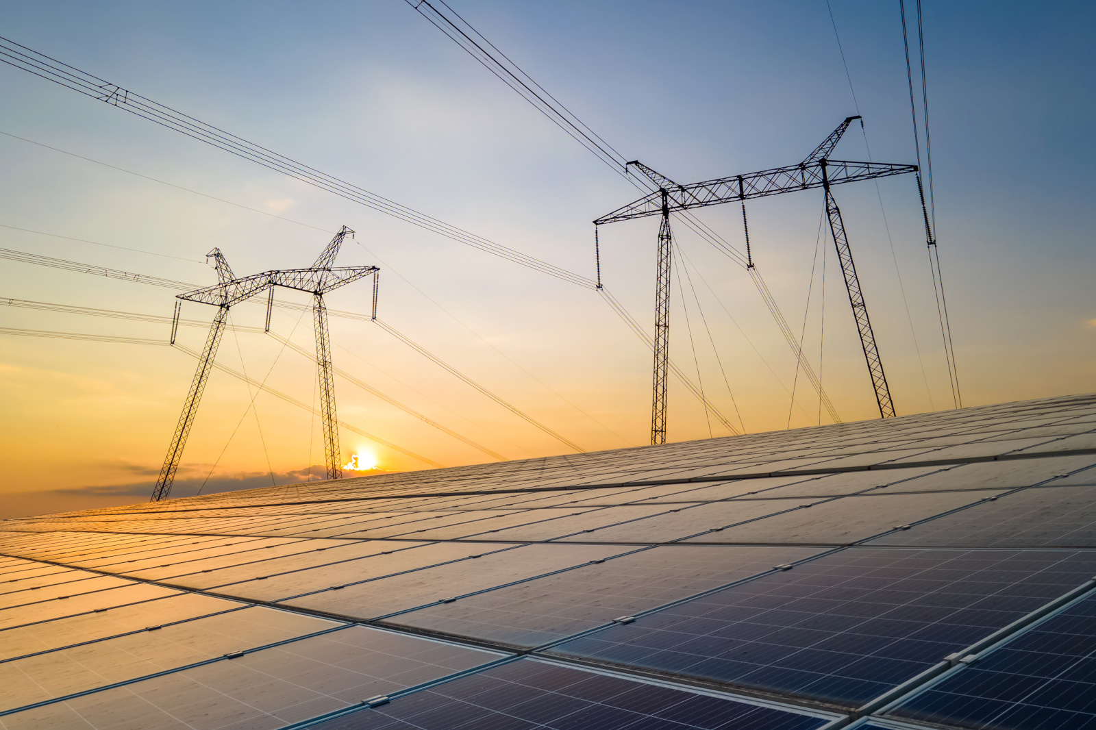 Zonnepanelen zijn geïnstalleerd onder hoogspanningsleidingen tegen de achtergrond van een zonsondergang.