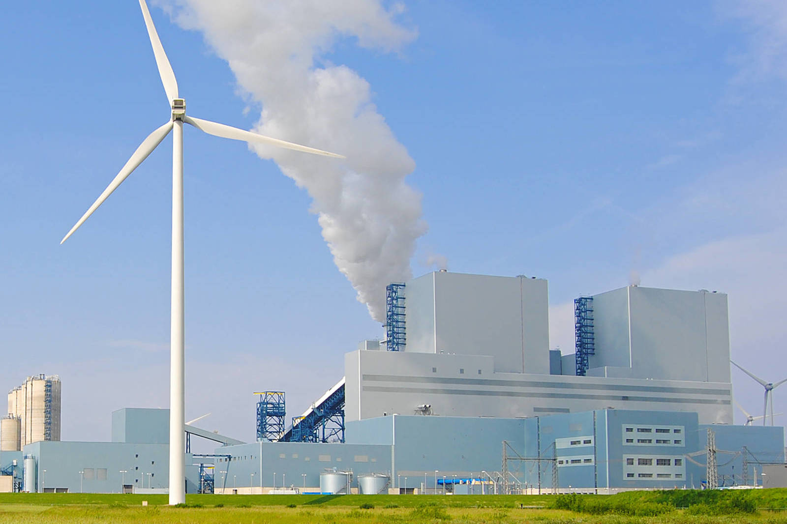 Een windturbine staat naast een grote industriële installatie die rook in de blauwe lucht uitstoot.