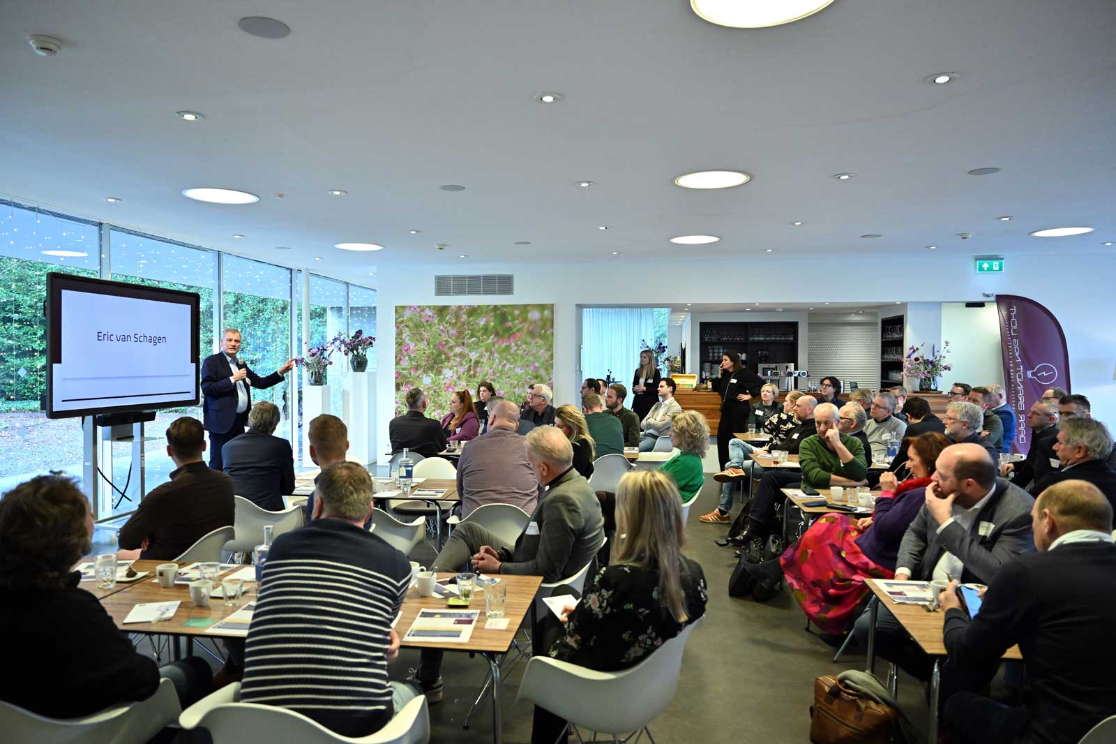 A business seminar with attendees focused on a speaker, Eric van Schagen, presenting at a modern venue with large windows.