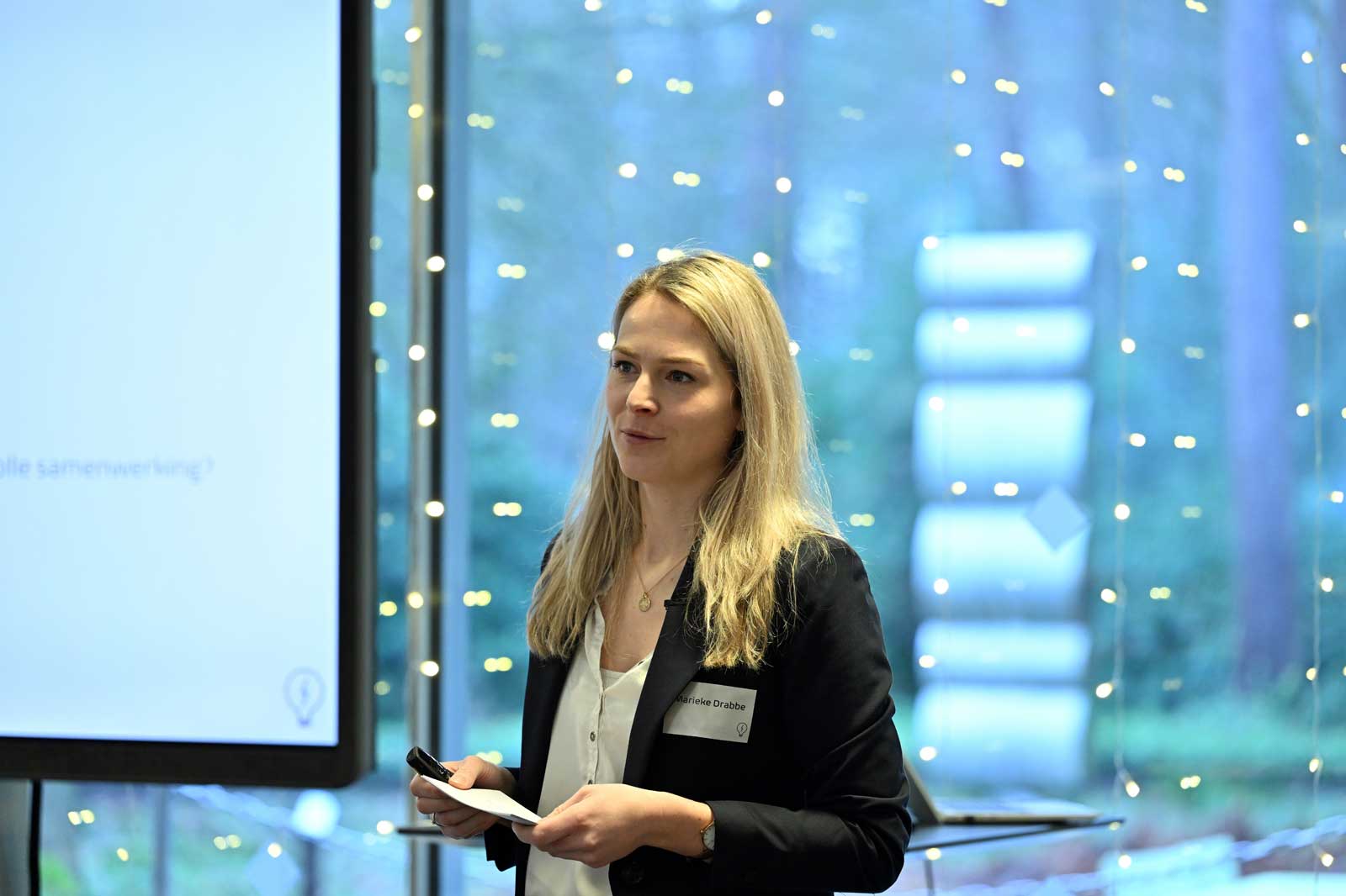 A woman presenting with a notepad in a well-lit space decorated with fairy lights and a large screen behind her.
