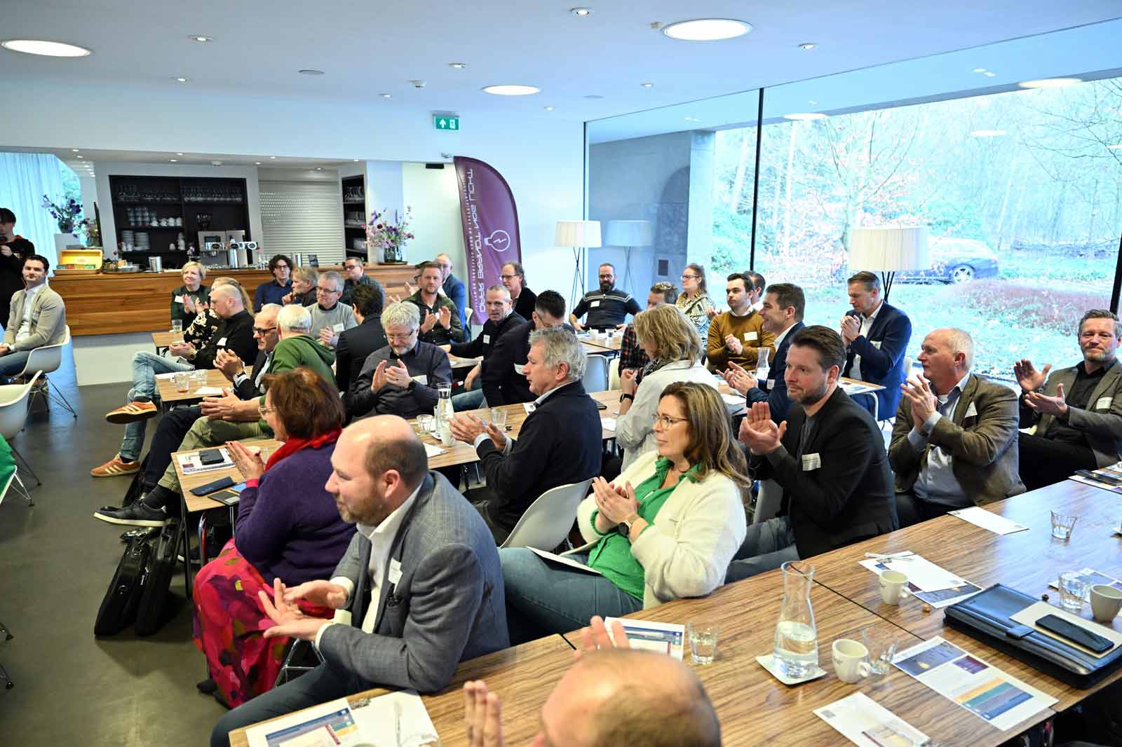 Een diverse groep mensen die applaudisseren in een moderne vergaderruimte, met een houten tafel en grote ramen met uitzicht op het bos.