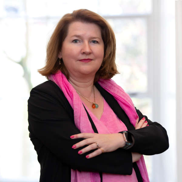 Een persoon staat met gekruiste armen. Ze dragen een roze top en sjaal tegen een heldere achtergrond.