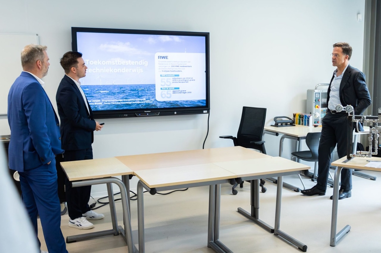 Three men in a modern classroom discuss a presentation displayed on a large screen, featuring graphs and information about RWE.