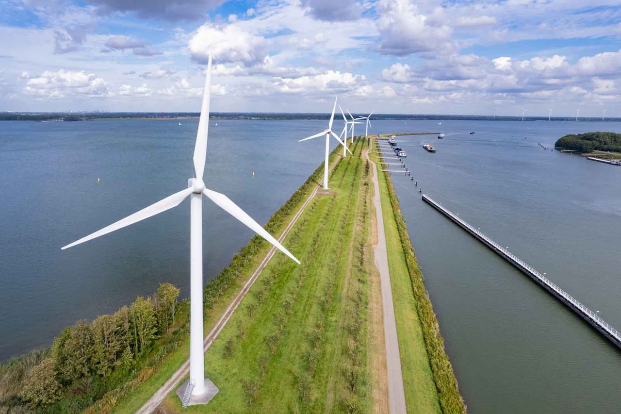 Aerial view of wind turbines along a green path beside a calm body of water under a partly cloudy sky.