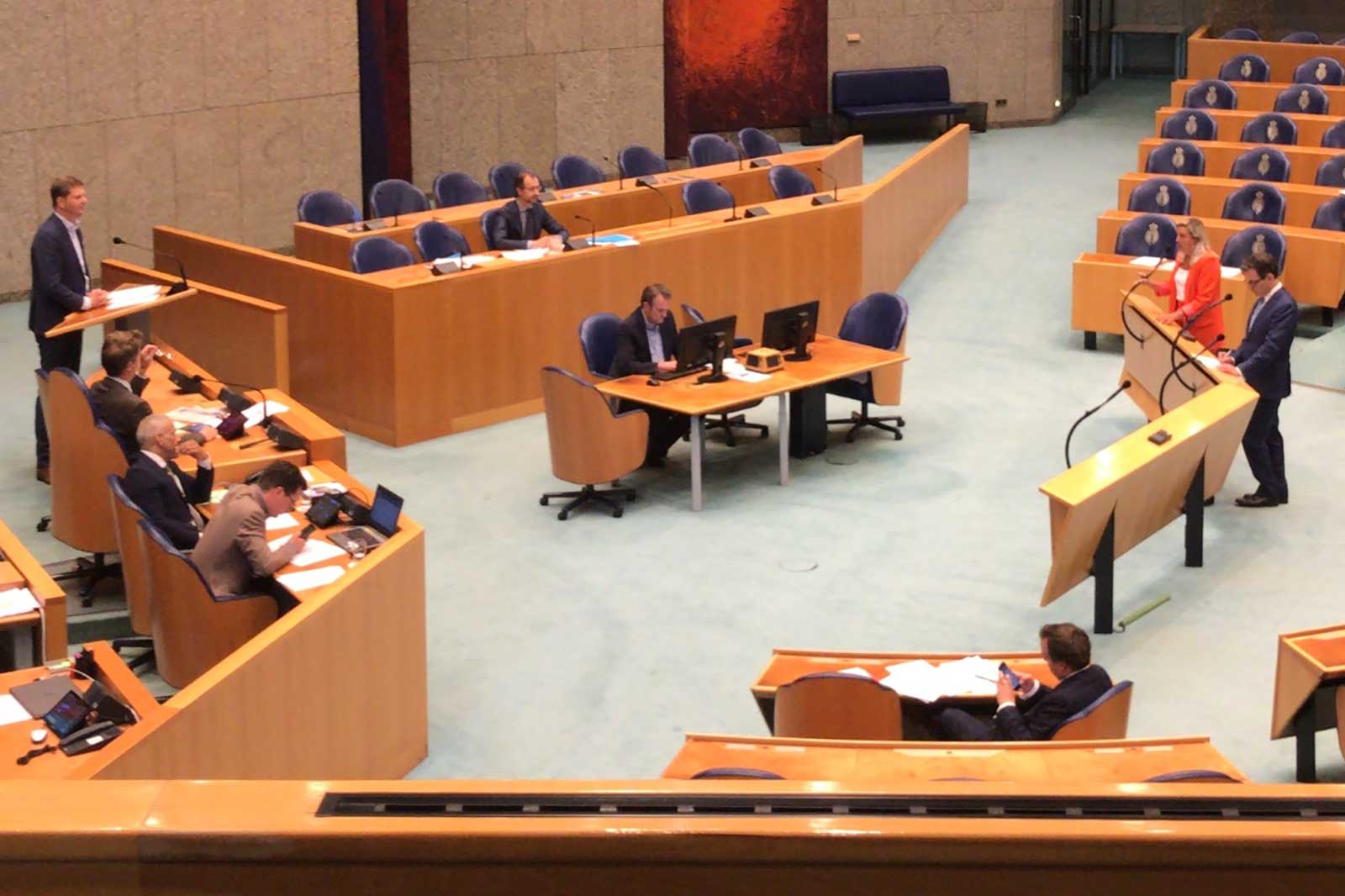 Een parlementaire sessie in uitvoering met meerdere deelnemers achter bureaus en een spreker op een spreekgestoelte.