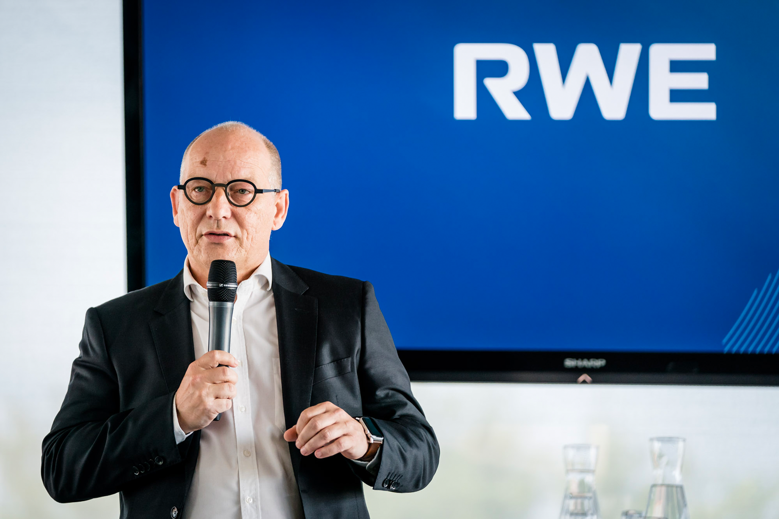 Een spreker in een zwart pak houdt een microfoon vast voor een groot scherm met het RWE-logo.