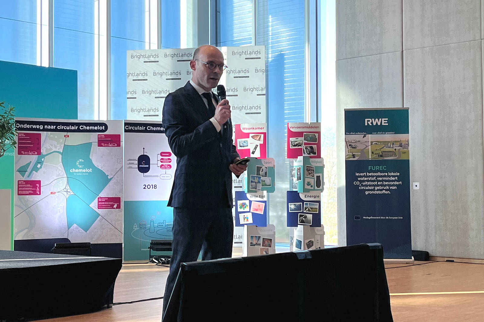 Een spreker in een pak presenteert op een conferentie, met visuele hulpmiddelen en banners op de achtergrond over circulaire projecten.