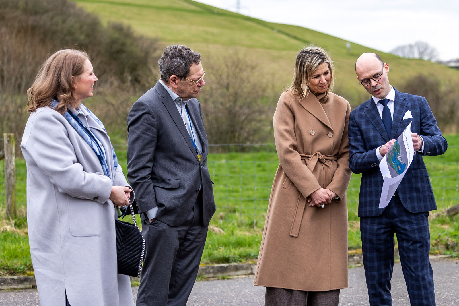 Vier mensen zijn buiten met elkaar in gesprek, met groene heuvels op de achtergrond en een man die een document vasthoudt.