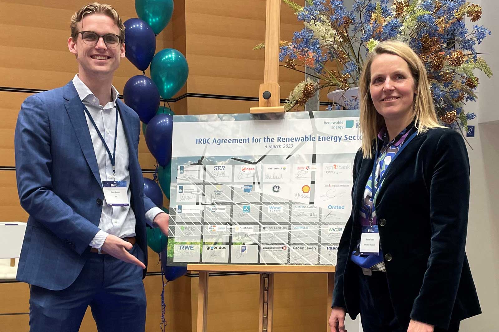 Twee personen staan naast een poster over de IRBC-overeenkomst voor de duurzame energiesector, omringd door ballonnen.